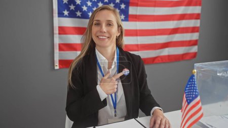 A smiling young caucasian woman points to her 'i voted' sticker in an american indoor voting center with flags.