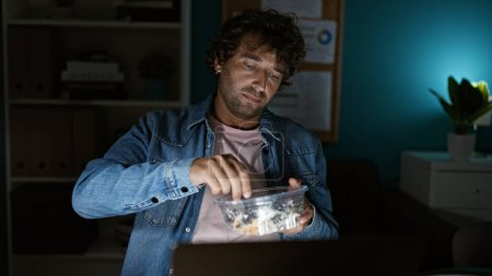 A young hispanic man in a casual denim jacket eats salad during a late night at the office.