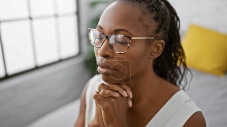 Eine nachdenkliche afrikanisch-amerikanische Frau mit lockigem Haar und Brille betet zu Hause