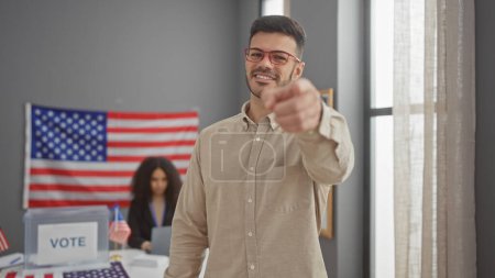Selbstbewusster Mann zeigt drinnen auf ein US-Wahlzentrum mit einer Frau und amerikanischen Flaggen im Hintergrund.