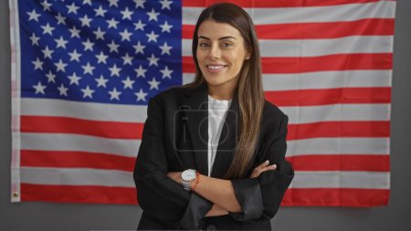 Téléchargez les photos : Femme hispanique en tenue professionnelle avec les bras croisés devant un drapeau américain à l'intérieur. - en image libre de droit