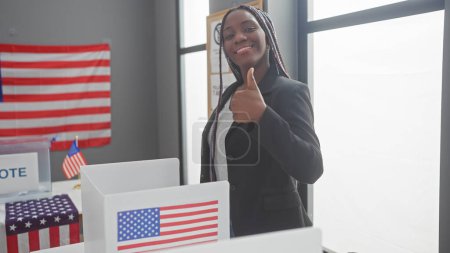 Una mujer afroamericana con trenzas da un pulgar hacia arriba dentro de un centro de votación adornado con banderas de estados unidos.