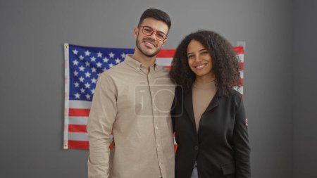 Ein Mann und eine Frau in professioneller Kleidung lächeln in einem Büro, im Hintergrund die amerikanische Flagge