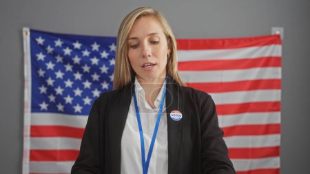 A young blonde woman with a 'voted' sticker, posing in front of an american flag indoors, symbolizing us electoral participation.