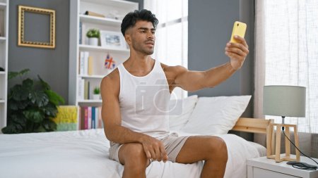 Ein hübscher junger hispanischer Mann in weißem Tank-Top macht ein Selfie in einem modernen Schlafzimmer mit grauer Wand.
