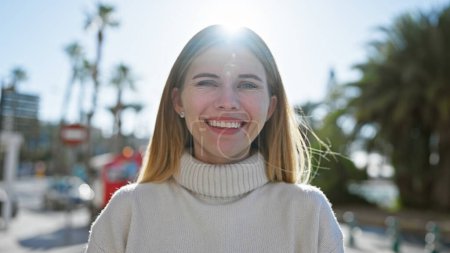 Téléchargez les photos : Une femme blonde joyeuse aux yeux bleus portant un pull blanc pose dans un cadre urbain ensoleillé. - en image libre de droit
