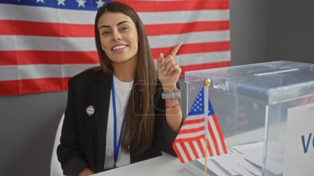 Foto de Sonriente joven hispana señalando mientras está en el centro electoral americano, con urnas y bandera. - Imagen libre de derechos