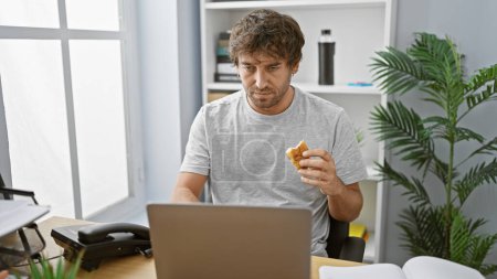 Photo for A bearded man in casual attire takes a snack break at his modern office workspace, exuding a relaxed atmosphere. - Royalty Free Image