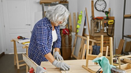 Une femme mûre travaille attentivement dans un atelier de menuiserie, entourée de meubles et d'outils en bois.