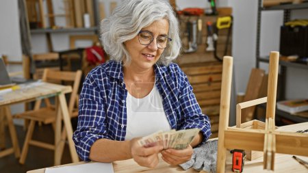 Foto de Una mujer de pelo gris cuenta con cuentas de zloty pulido en un taller de carpintería, rodeado de herramientas de carpintería. - Imagen libre de derechos