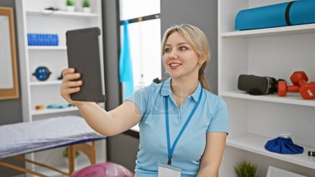 Photo for Blonde woman in healthcare uniform taking a selfie with smartphone at a modern rehab clinic. - Royalty Free Image