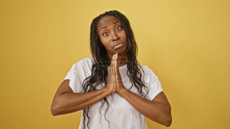 Adult african american woman with curly hair, pleading hands, isolated on a yellow background.