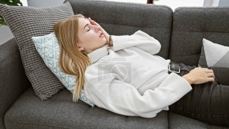 Téléchargez les photos : Femme blonde reposant confortablement sur un canapé gris dans un salon confortable, respirant une ambiance sereine et chaleureuse. - en image libre de droit