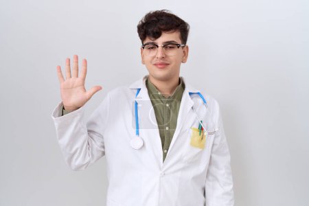Foto de Young non binary man wearing doctor uniform and stethoscope showing and pointing up with fingers number five while smiling confident and happy. - Imagen libre de derechos
