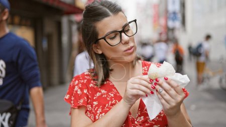 Schöne hispanische Frau genießt ein köstliches Krabbenfleisch-Brötchen in der berühmten Dotonbori-Straße in Osaka, ihre Brille reflektiert die pulsierenden Lichter der Stadt