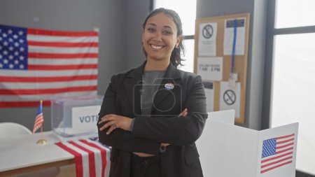 Lächelnde junge hispanische Frau mit verschränkten Armen steht in einem mit amerikanischen Flaggen und einer Wahlkabine dekorierten Raum, die auf eine Wahlkulisse in den Vereinigten Staaten hinweist.