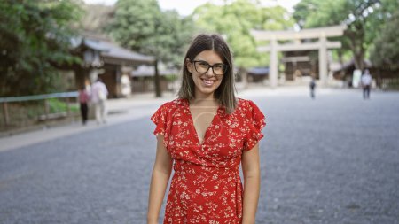 Cheerful, beautiful hispanic woman with glasses poses confidently, smiling at tokyo's meiji shrine, her joy unequivocally radiating through her astonishing smile