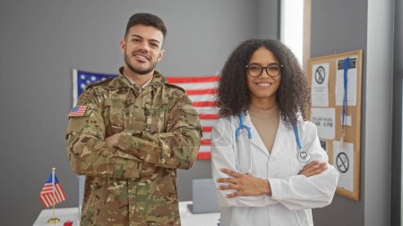 Photo for A man in military uniform and a woman doctor standing with arms crossed in front of a usa flag, indoors. - Royalty Free Image