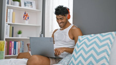 A cheerful hispanic man with beard wearing headphones enjoys using his laptop in a cozy bedroom setting.