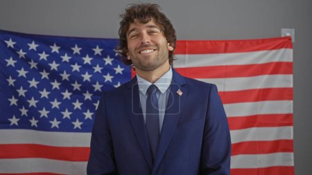Foto de Un joven hispano adulto sonriente con barba con traje se para ante una bandera americana dentro de una oficina. - Imagen libre de derechos