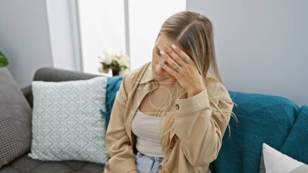 Mujer rubia joven estresada sentada sola en el sofá en casa, luchando con dolor de cabeza y emoción en una batalla con la depresión.
