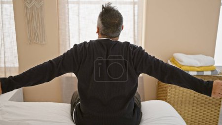 Ein reifer hispanischer Mann mit grauen Haaren und Bart streckt sich in seinem Schlafzimmer und beginnt seine morgendliche Routine.