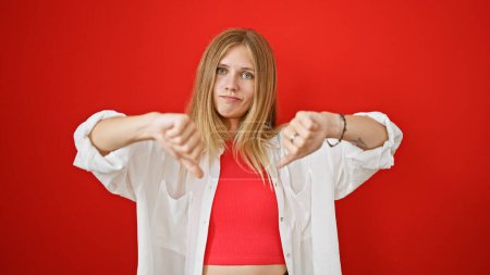 A skeptical young woman with blonde hair gives a thumbs down gesture against a vibrant red background.
