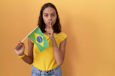 Téléchargez les photos : Jeune femme hispanique tenant le drapeau brésilien demandant à se taire avec le doigt sur les lèvres. silence et concept secret. - en image libre de droit