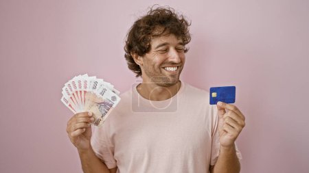 Guapo joven sonriendo mientras sostiene coronas icelándicas y tarjeta de crédito sobre un fondo rosa