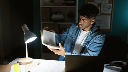 Hispanic man reading notebook in a dimly lit office at night.