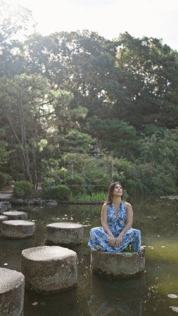 Beautiful hispanic woman sitting on a stone path, taking in nature's green summer splendor by a serene lake at heian jingu, kyoto - a divine journey, looking around traditional japanese temple