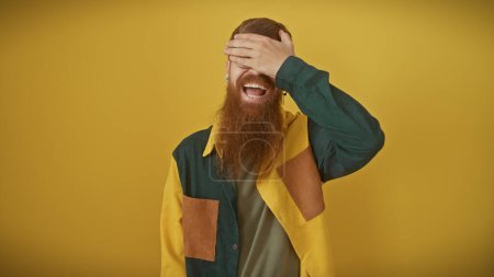 A cheerful, young, redhead man with a beard, laughing while covering his eyes, stands against an isolated yellow background.