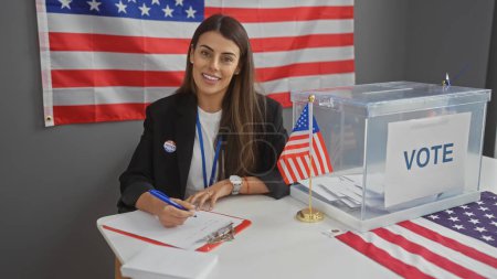 Una joven hispana sonriente se sienta en el interior con una bandera americana, tomando notas en un centro de votación del colegio electoral.