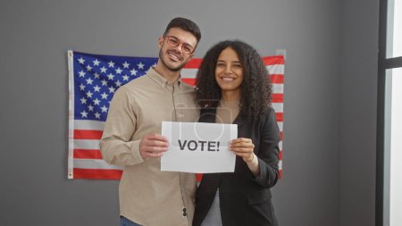 Smiling man and woman holding a 'vote!' sign in front of an american flag inside an office.