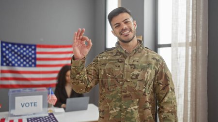 Foto de Un soldado sonriente en camuflaje haciendo un gesto de "ok" en el interior, con una mujer y nosotros bandera en el fondo en un centro de votación - Imagen libre de derechos