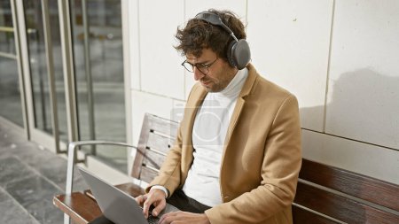 Photo for Handsome young hispanic man with beard and headphones using laptop on city street bench. - Royalty Free Image