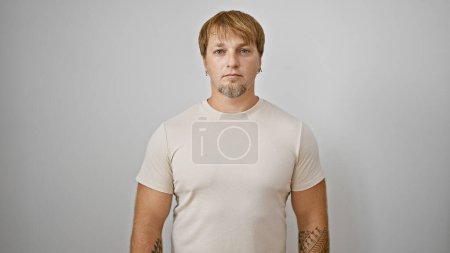 Portrait of a young adult man with blond hair and beard, tattooed arms, wearing a beige t-shirt, against a white background.