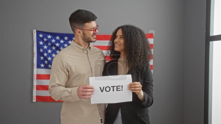 A man and a woman in professional attire stand indoors with a usa flag, holding a sign that says 'vote!'