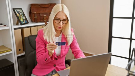 Téléchargez les photos : Jolie jeune femme blonde, une travailleuse d'affaires détendue mais sérieuse, payant en ligne avec une carte de crédit tout en travaillant sur un ordinateur portable à l'intérieur du bureau - en image libre de droit