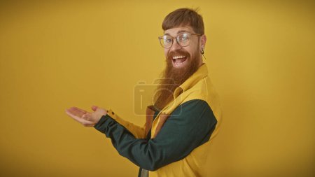 A cheerful bearded man in casual attire presents with an open hand against a solid yellow background