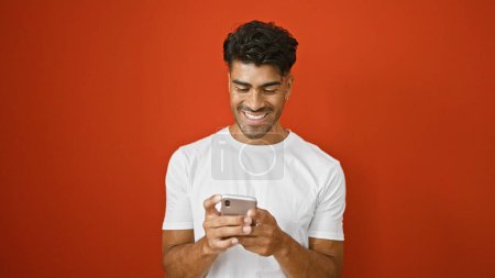 Photo for Handsome young hispanic man in white shirt smiling at phone against isolated red background - Royalty Free Image