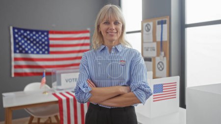 Foto de Una mujer rubia confiada con una pegatina 'votada' se para con los brazos cruzados en una sala de colegio electoral estadounidense con banderas - Imagen libre de derechos