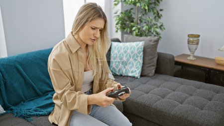 Attraktive junge blonde Frau, vertieft in eine intensive Online-Gaming-Session, bequem auf ihrem heimischen Sofa sitzend, völlig verloren in der Welt der digitalen Technologie und virtuellen Joystick-Steuerung.