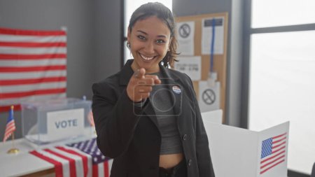 Foto de Mujer hispana señalando mientras sonríe en un centro electoral americano con cabinas de votación y banderas. - Imagen libre de derechos