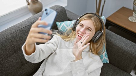 A cheerful blonde woman wearing headphones video calling from a cozy living room couch