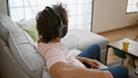 Photo for A relaxed man with headphones sits on a couch in a modern living room, enjoying leisure time. - Royalty Free Image