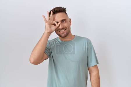 Photo for Hispanic man with beard standing over white background doing ok gesture with hand smiling, eye looking through fingers with happy face. - Royalty Free Image