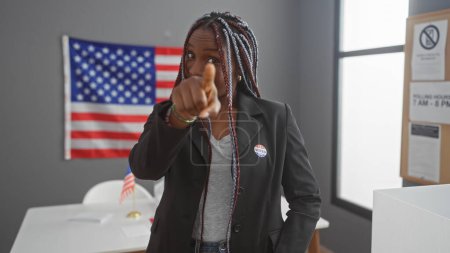 Una mujer afroamericana confiada con trenzas apuntando a la cámara en un centro de votación universitario con una bandera americana en el fondo.