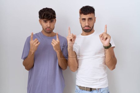 Couple homosexuel gay debout sur fond blanc pointant vers le haut regardant triste et contrarié, indiquant la direction avec les doigts, malheureux et déprimé. 
