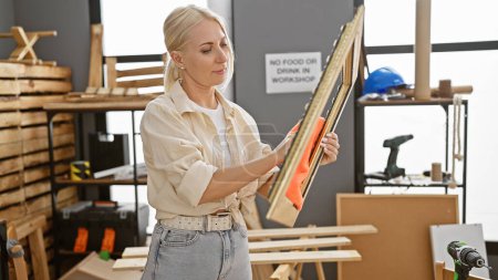 Téléchargez les photos : Jolie et jeune femme blonde menuisière qui nettoie le cadre en bois propre à l'atelier de menuiserie, portrait de l'artisanat industriel du bois - en image libre de droit